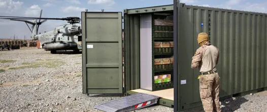 Organized and Secure: Field Storage for Military Inventory and Supplies Olive drab container open in a desert field with shelves of supplies; a soldier and large military helicopter are nearby.