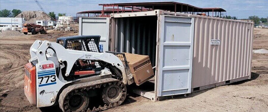 Durable On-Site Storage: Shipping Containers for Farm Equipment and Tools Skid-steer loading large equipment into an open tan shipping container on a dirt construction or agricultural site.