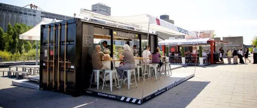 High-Traffic Retail: Container Bars, Cafés, and Outdoor Food Kiosks Black shipping container restaurant with a fold-down patio deck, awning, and customers seated at a bar in a sunny urban setting.