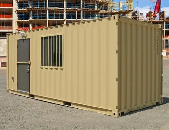 Tan modified shipping container with a secure door and barred window, used for tool and material storage at a construction site.