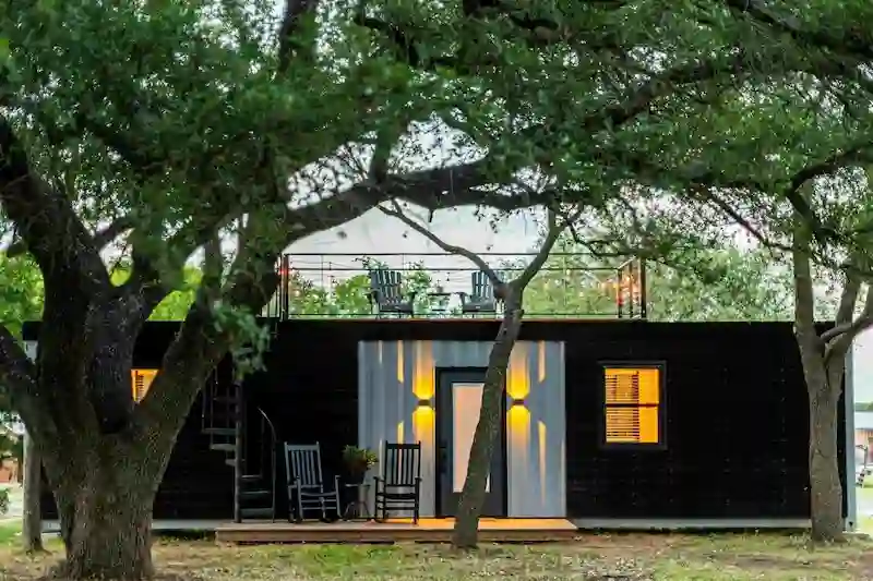 Black container home with a front porch, rocking chairs, and a spiral staircase leading to a rooftop deck.