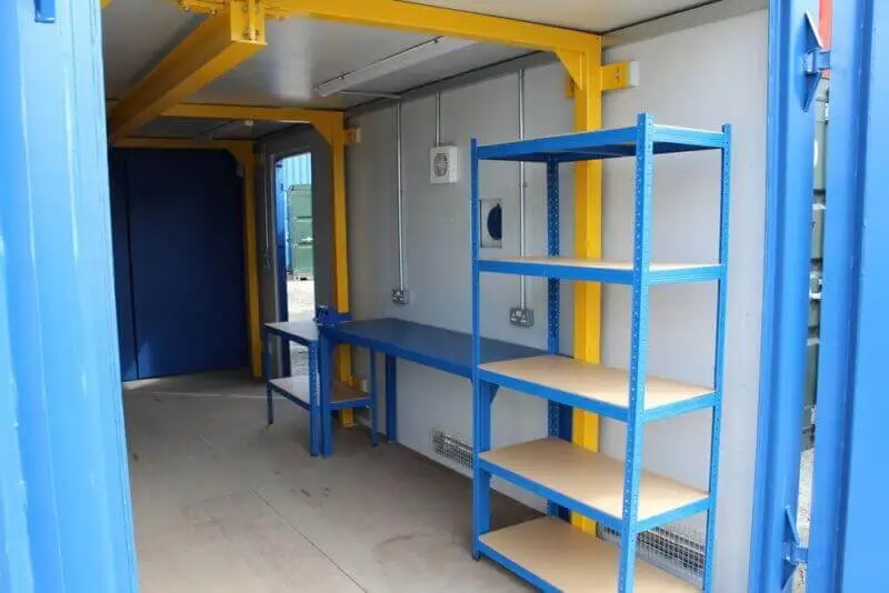 Interior de un taller en un contenedor de transporte modificado, mostrando estanterías azules, un banco de trabajo, tomas eléctricas y vigas estructurales amarillas.