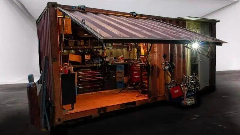 Contenedor oxidado muy modificado, con el lateral plegado en forma de toldo, que revela un completo taller de soldadura en su interior.