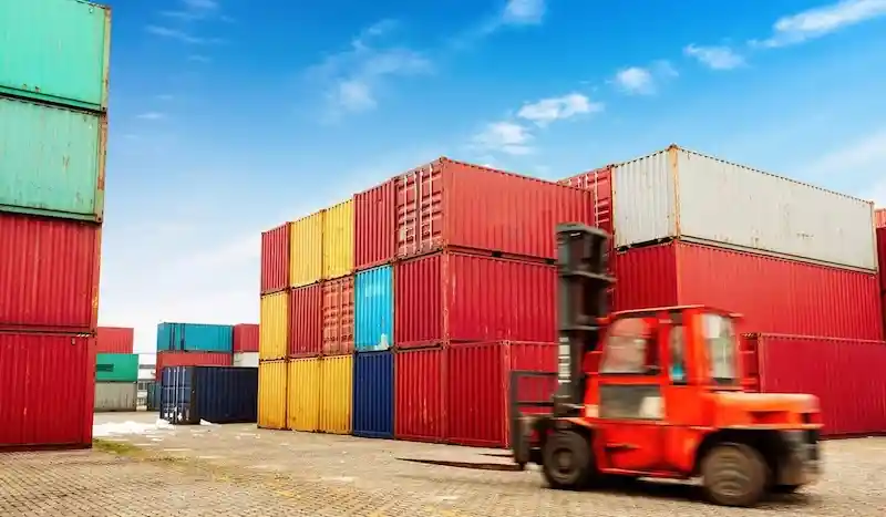 Carretilla elevadora roja moviéndose rápidamente entre grandes pilas de contenedores de transporte multicolores bajo un cielo brillante.