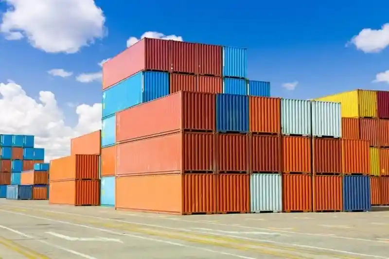Coloridas pilas de contenedores de transporte rojos, naranjas, azules y amarillos bajo un cielo azul brillante, que simbolizan un vasto inventario.