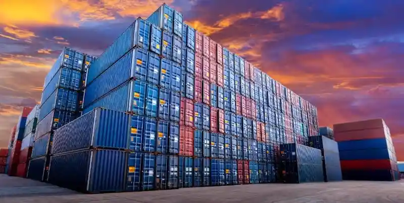 Dramática foto al atardecer de imponentes pilas de contenedores de transporte marítimo azules y rojos, que simbolizan el enorme peso de la logística.
