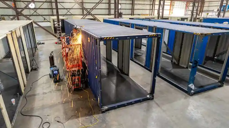 Welder working on a blue shipping container frame with sparks flying in a factory.