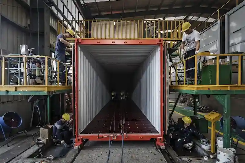 Factory workers in hard hats finishing the interior of a red shipping container.