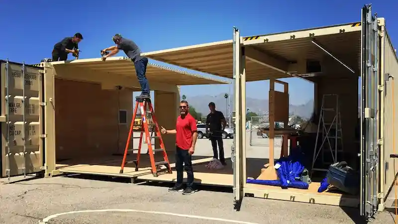 Three workers modifying a beige shipping container with a fold-out side panel outdoors.