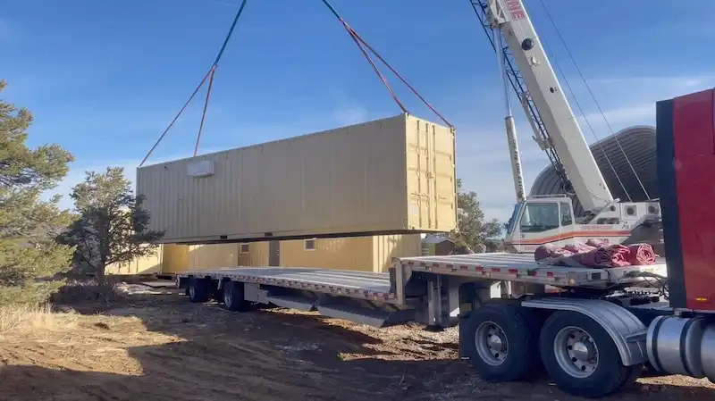 Camión grúa grande levantando un contenedor de transporte beige de su remolque de plataforma para colocarlo con precisión en una propiedad rural.