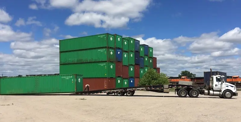 Camión semirremolque blanco utilizando un remolque de plataforma basculante para dejar caer un gran contenedor de transporte verde en un solar de tierra cerca de un inventario apilado.