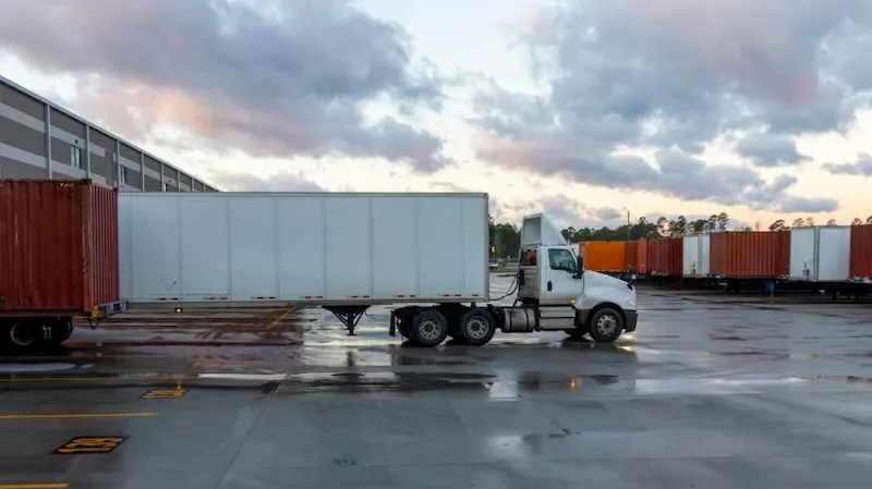 Semirremolque blanco tirando de un remolque en un patio de depósito húmedo al anochecer, rodeado de contenedores de transporte apilados.