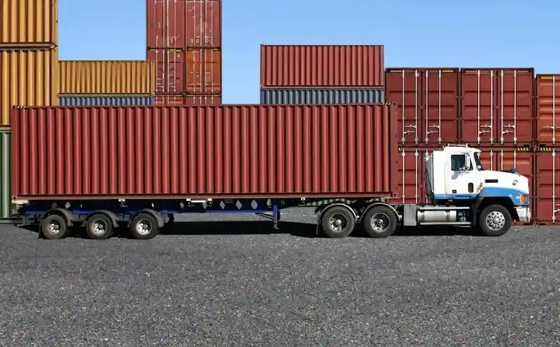 Camión semirremolque con remolque de plataforma que transporta un largo contenedor rojo, con otros contenedores apilados al fondo.