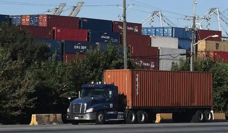Camión azul que transporta un gran contenedor naranja, pasando junto a densas pilas de contenedores multicolores y grúas en un puerto.
