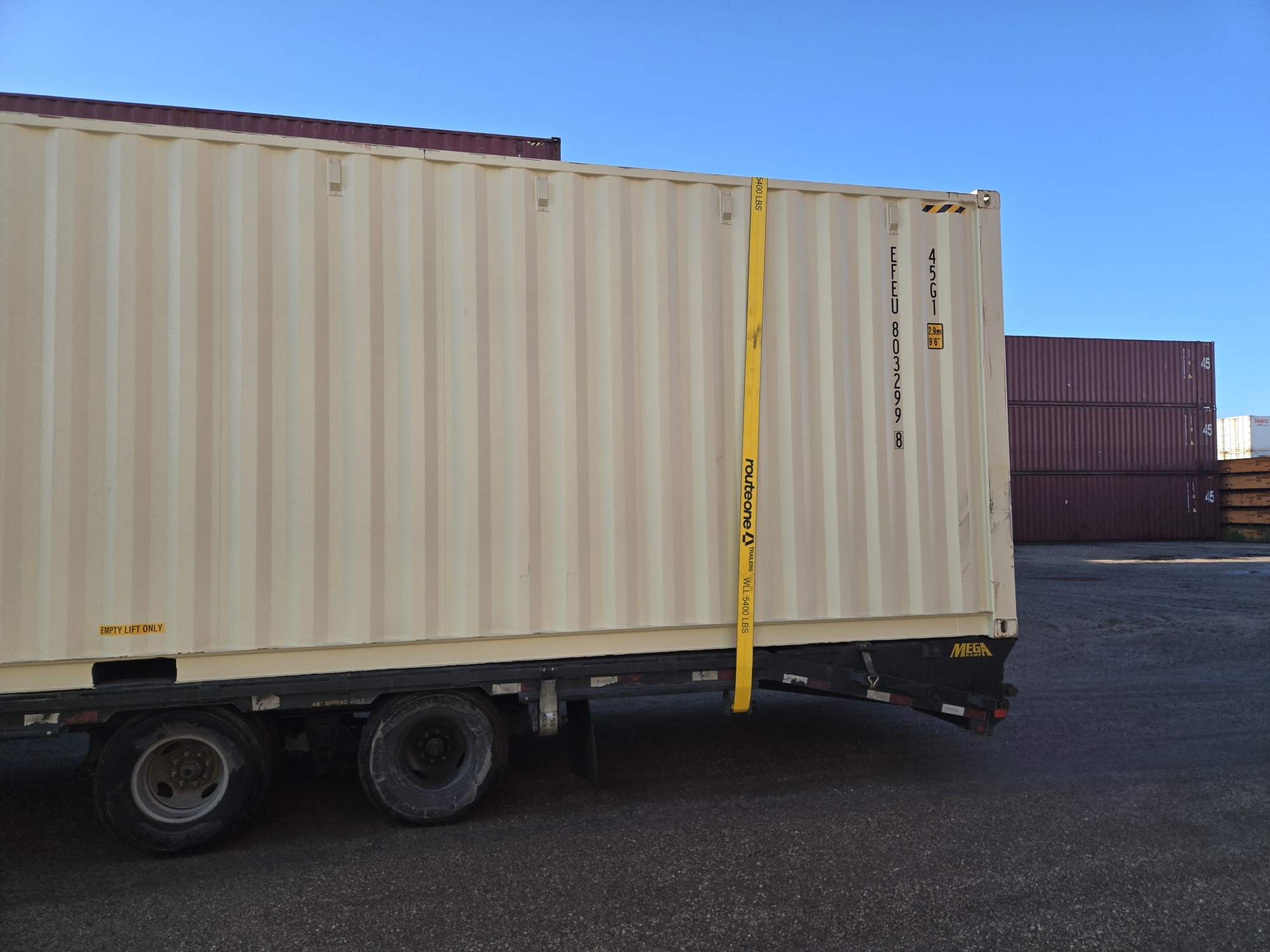 Side view of a 40ft high cube new shipping container secured on a tilt-bed trailer in a paved container yard