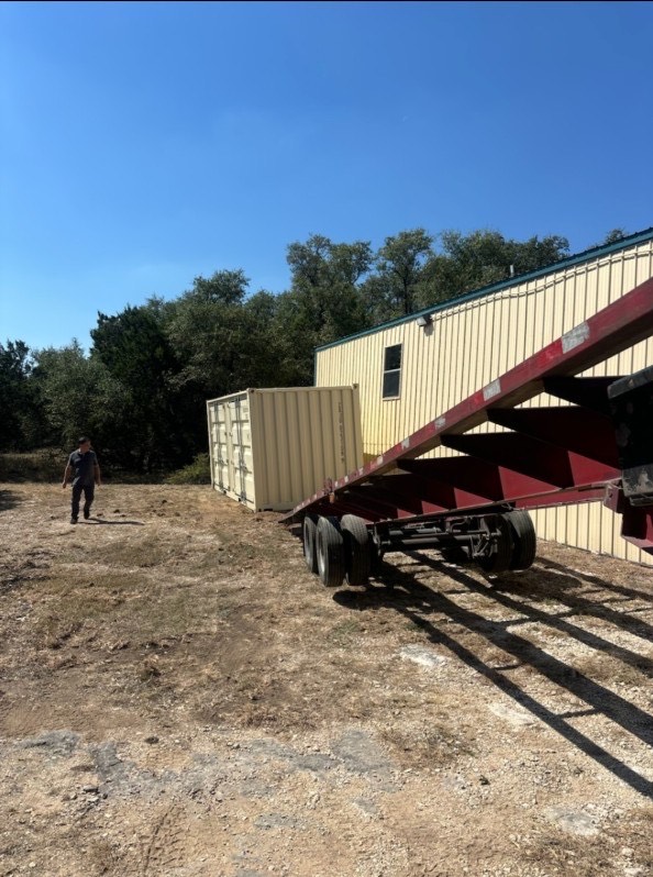 Red tilt-bed trailer sliding a beige 20ft container onto a gravel site