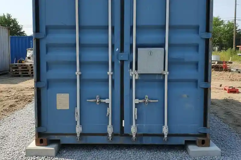 A blue used container door with a factory lockbox resting on concrete footings and a leveled gravel foundation.